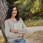 Mylène Muller, souriante, posée contre un pin, tenant son ouvrage CAP de créer la vie qui te ressemble ? 52 défis pour réenchanter ta vie